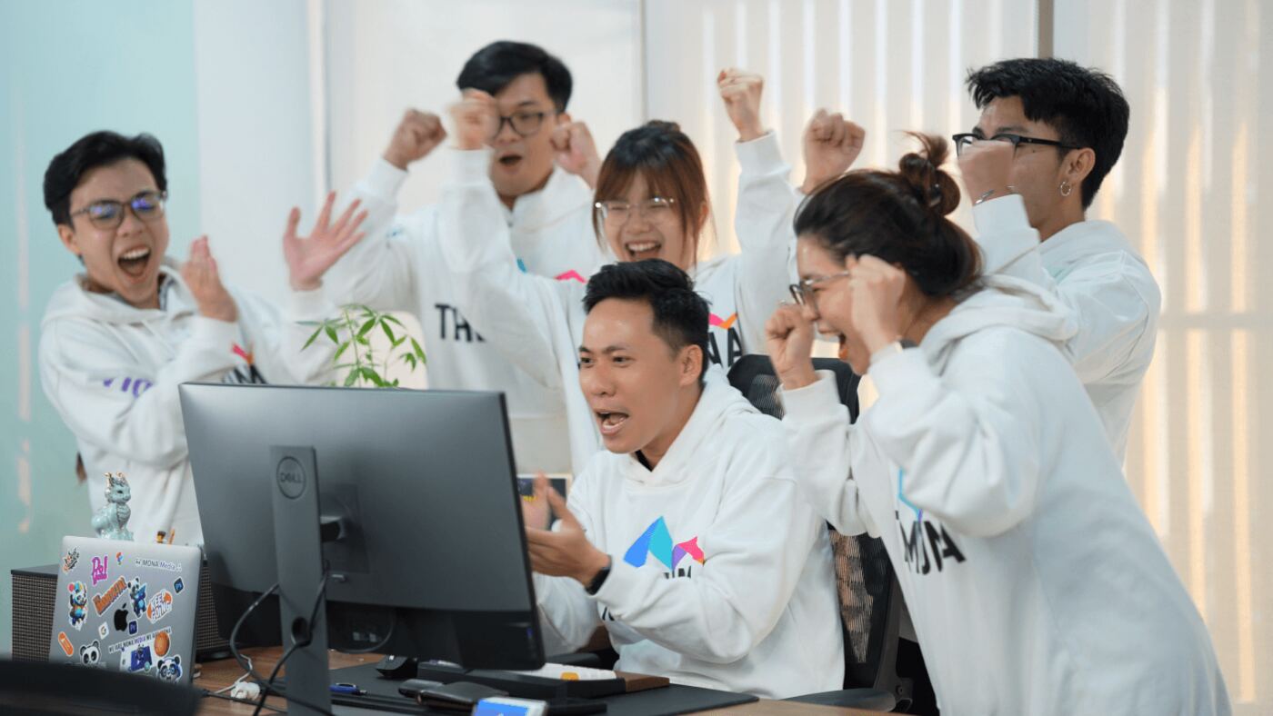 Nhân viên MONA mặc hoodie reo mừng trước màn hình máy tính tại văn phòng